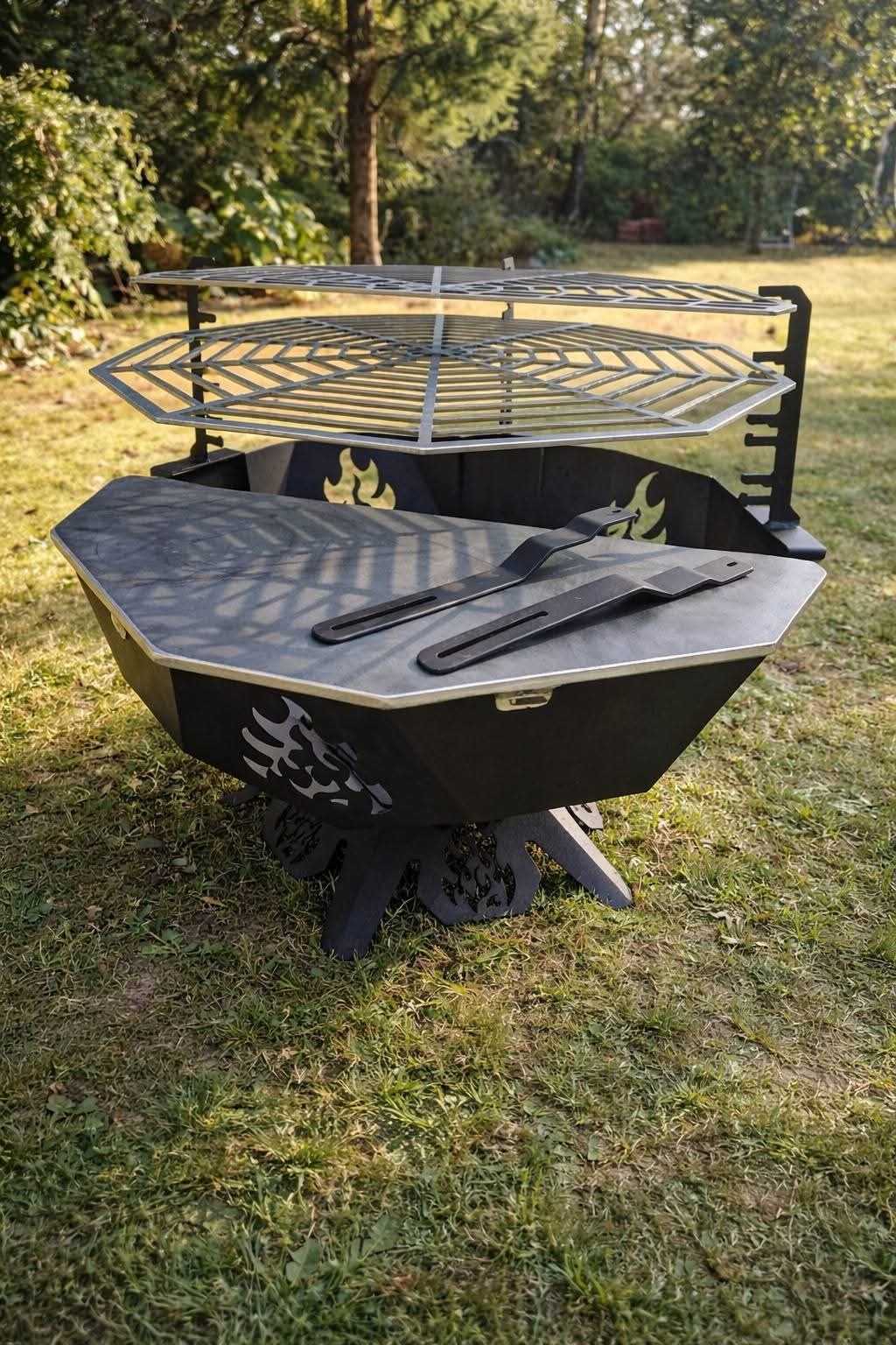 Czarny, metalowy grill ogrodowy z rusztem i blatem, zdobiony motywem ognia i smoka, ustawiony na trawie w ogrodzie. Widoczne narzędzia do grillowania.