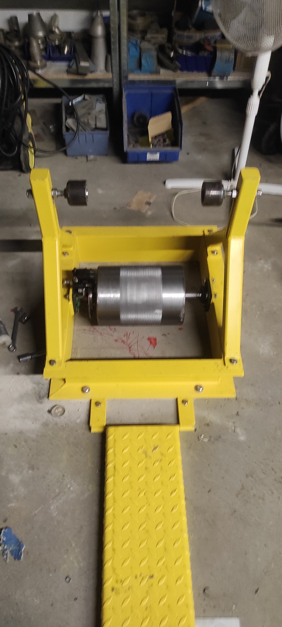 Żółta konstrukcja stalowa z zamontowanym silnikiem elektrycznym, widoczne elementy mocujące i narzędzia w tle. Detal z warsztatu ślusarskiego.