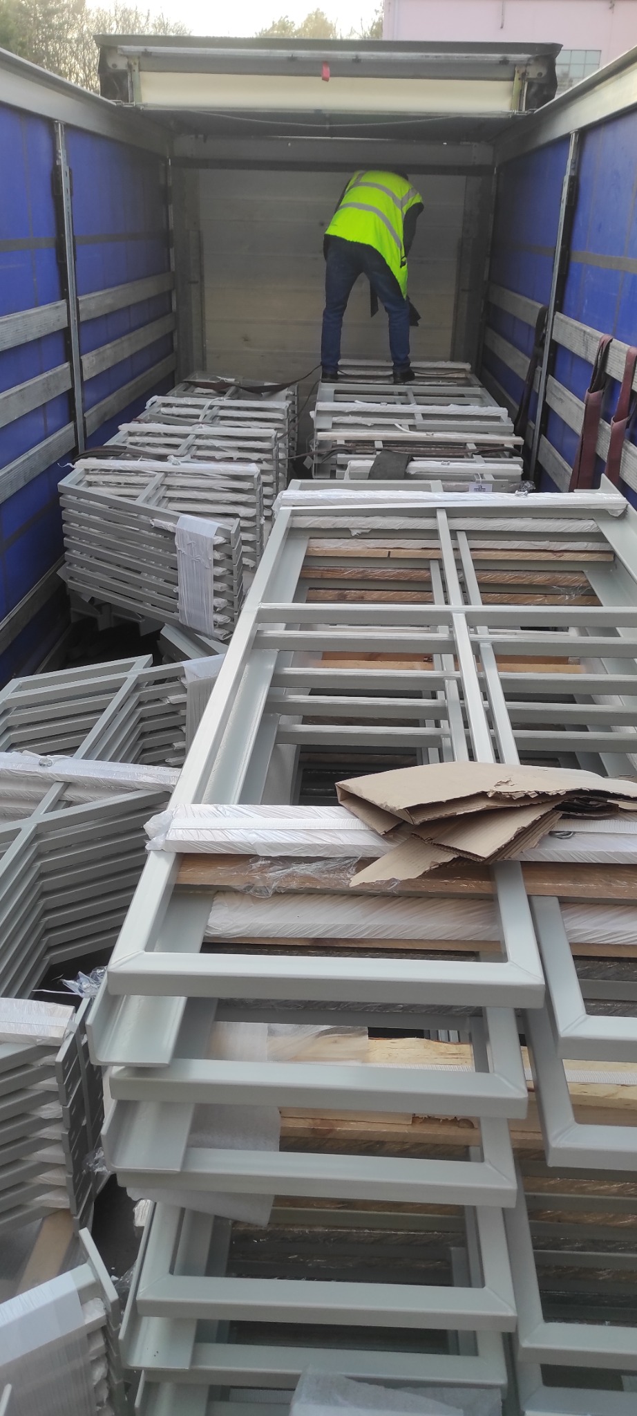 Transport metalowych ram konstrukcyjnych wewnątrz naczepy, zabezpieczonych folią i kartonem, z widoczną sylwetką osoby w odblaskowej kamizelce.