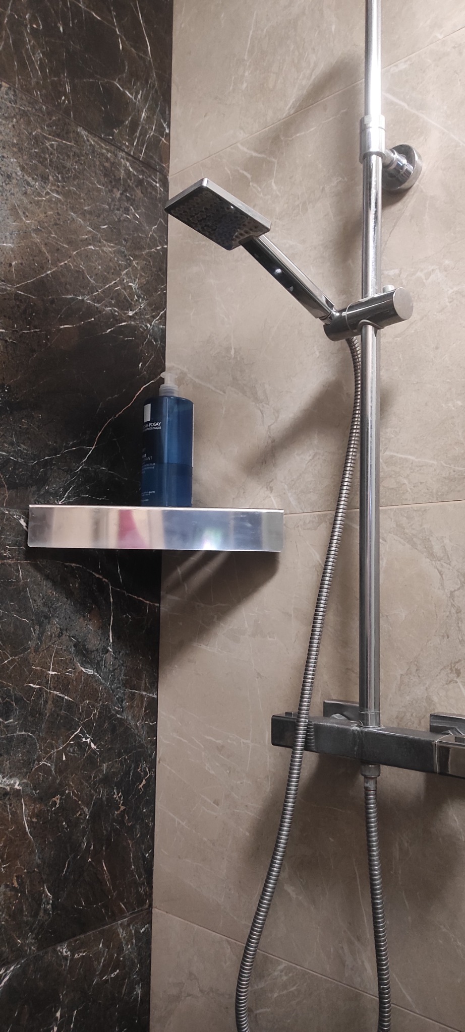 Nowoczesna kabina prysznicowa z chromowaną armaturą i półką, na której stoi butelka kosmetyku. Ściany wyłożone płytkami imitującymi marmur w odcieniach szarości.