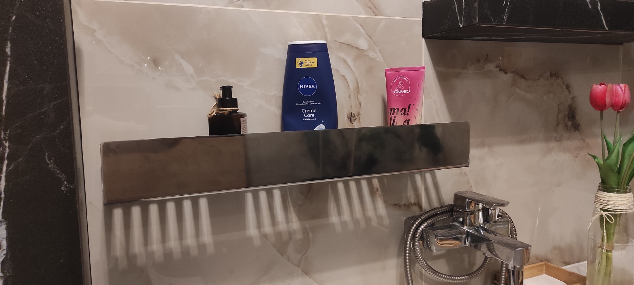 Półka łazienkowa ze stali nierdzewnej z kosmetykami na tle marmurowej ściany, obok bateria umywalkowa i wazon z tulipanami. Minimalistyczny design.
