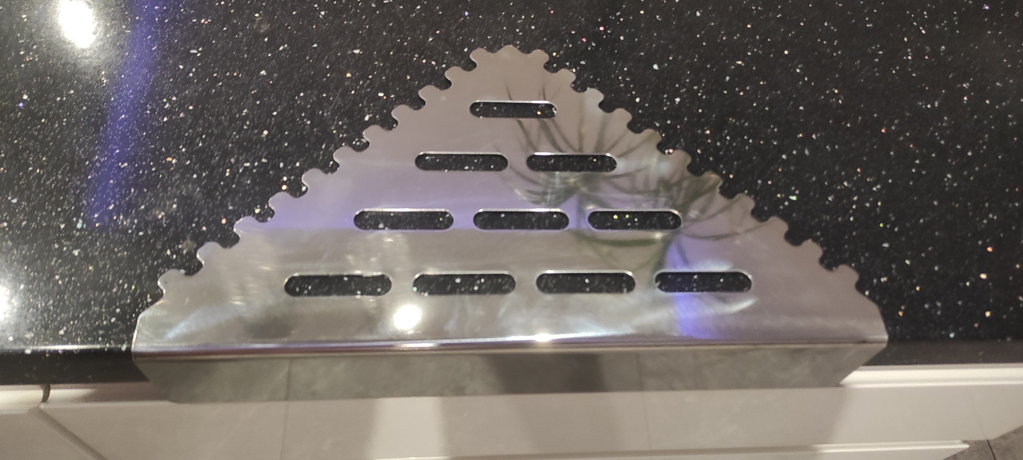 Metalowy, trójkątny element z ząbkowaną krawędzią i podłużnymi otworami, umieszczony na ciemnym blacie z połyskującymi drobinkami. Widoczne odbicie rośliny.