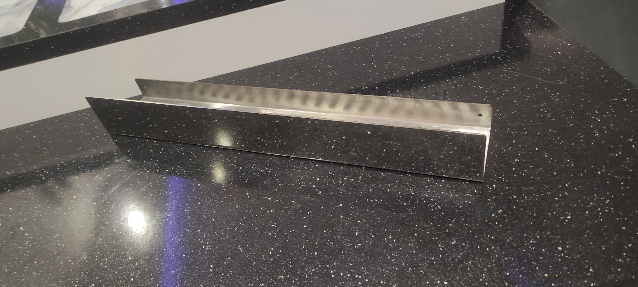 Nierdzewna, kątowa listwa przypodłogowa na blacie z czarnego, błyszczącego kamienia. Widoczne otwory montażowe. Minimalistyczny design.