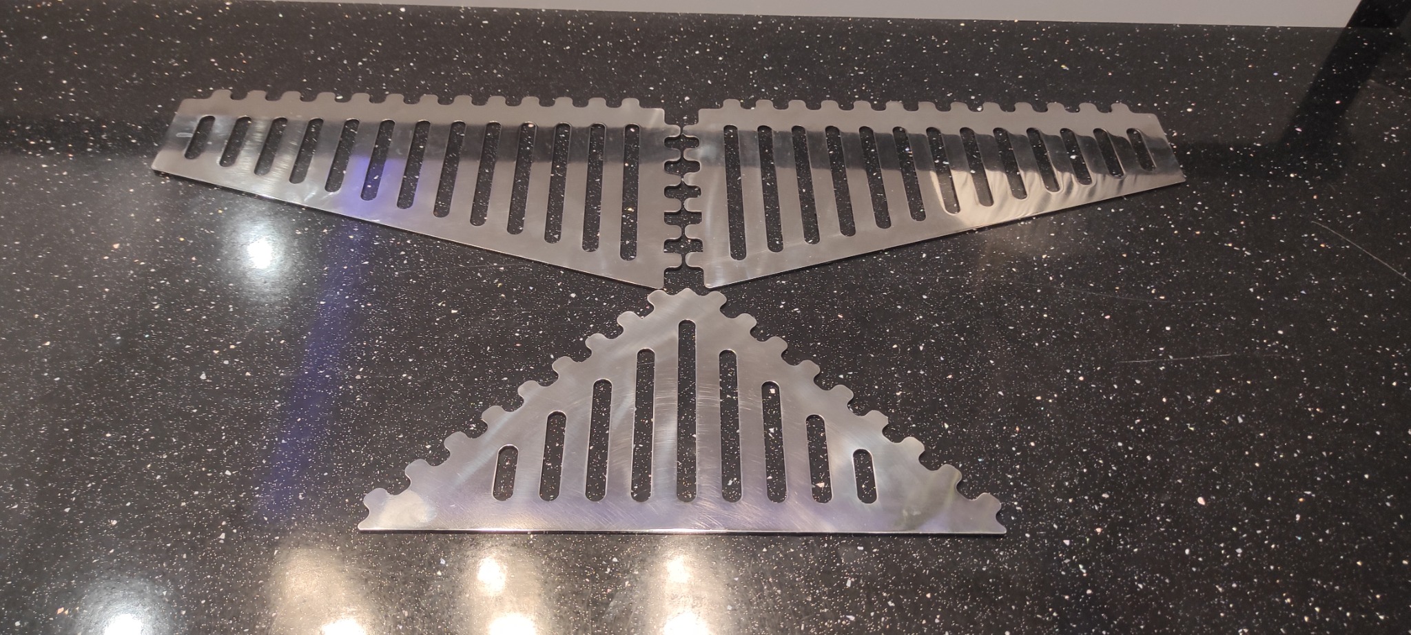 Metalowe elementy z wyciętymi otworami, prawdopodobnie do balustrady, leżą na blacie w Grodzisku Dolnym. Elementy są w kształcie trójkątów i prostokątów.