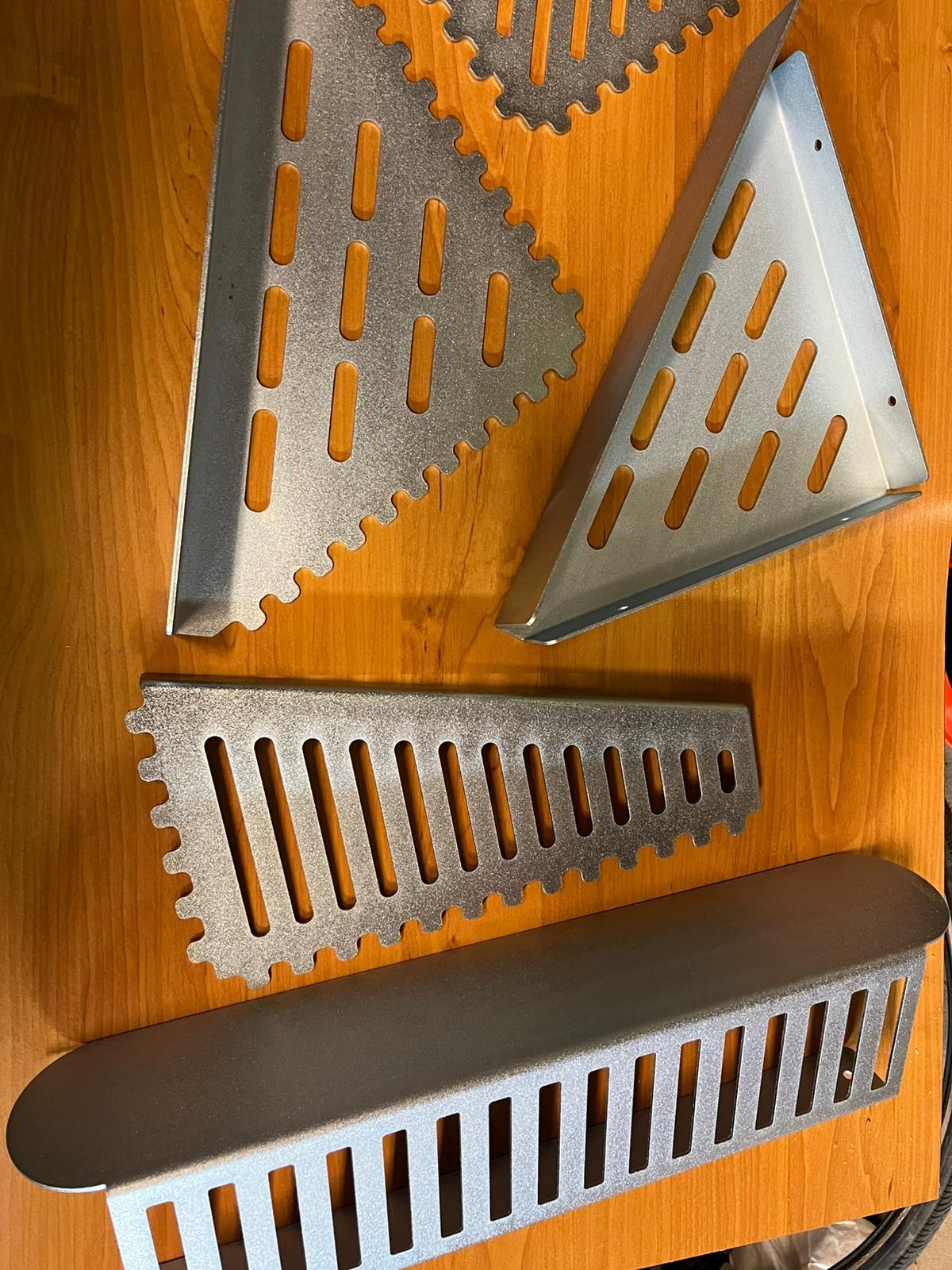 Metalowe elementy konstrukcyjne o różnych kształtach (trójkąt, prostokąt) z wyciętymi otworami, leżące na drewnianej powierzchni. Widoczne ząbkowane krawędzie.