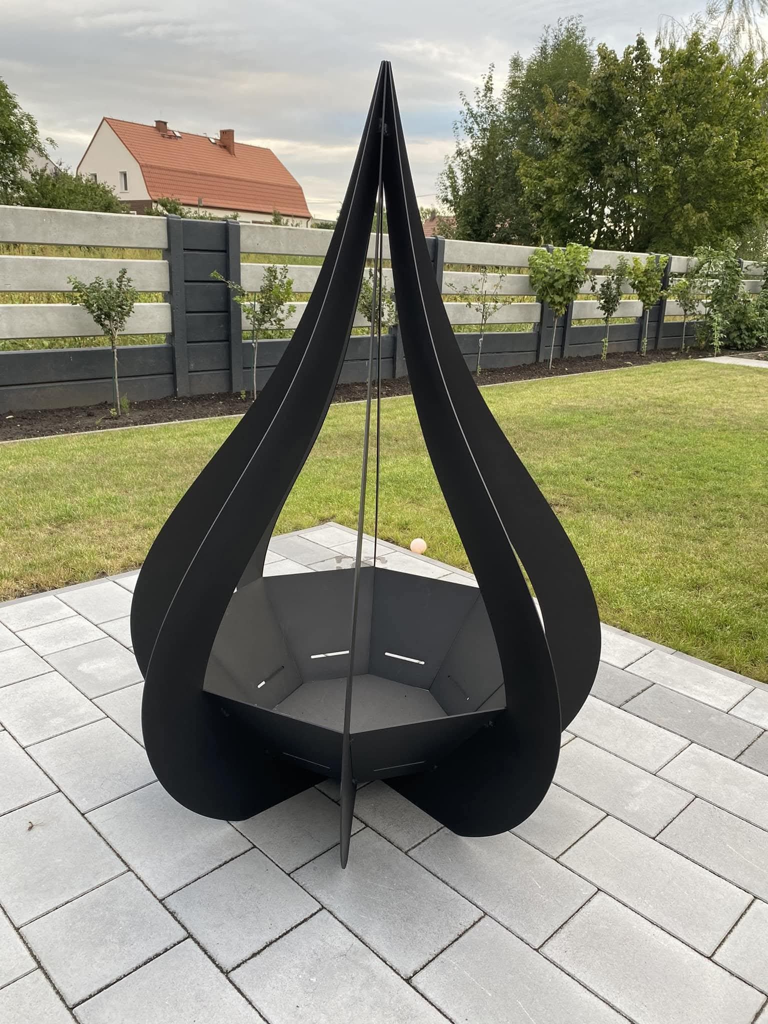 Czarny, metalowy kosz na ognisko o nowoczesnym designie, przypominającym płomień, stoi na szarym bruku w ogrodzie. W tle widoczne ogrodzenie i zieleń.