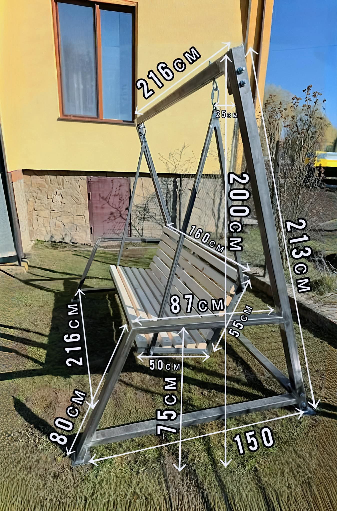 Metalowa huśtawka ogrodowa z drewnianym siedziskiem, z naniesionymi wymiarami, ustawiona na trawie przed budynkiem z elementami kamiennej elewacji.