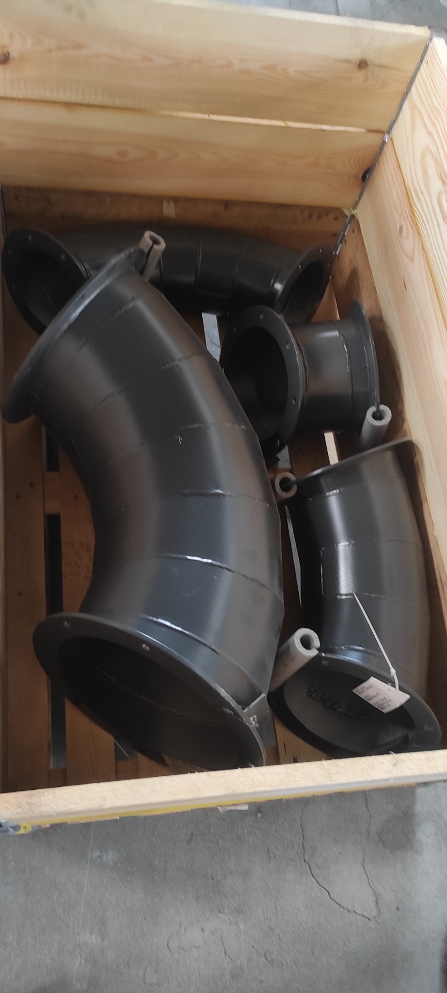 Czarne, metalowe kolanka z kołnierzami w drewnianej skrzyni. Elementy konstrukcji stalowej, prawdopodobnie do budowy hal lub instalacji przemysłowych. Widoczne spawy i etykiety.