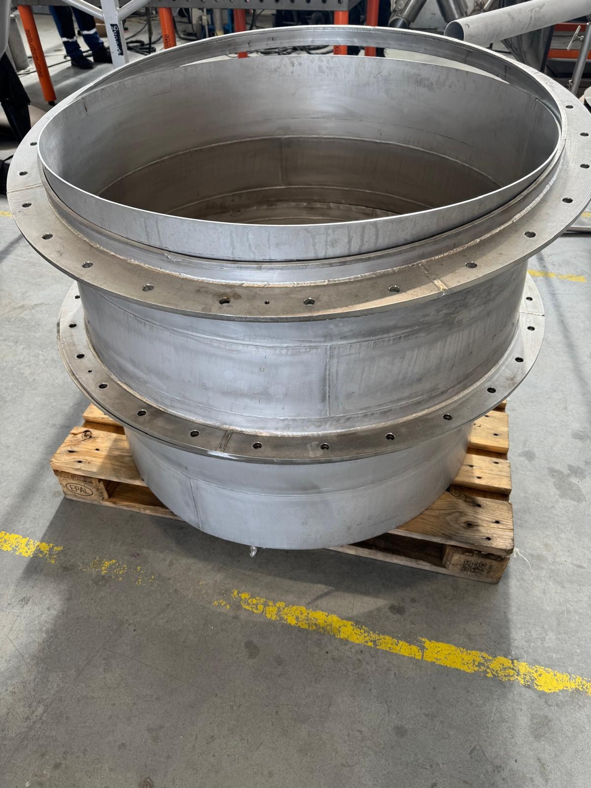 Element konstrukcyjny ze stali nierdzewnej, spawany, z otworami na śruby, umieszczony na drewnianej palecie. Widoczne ślady spawania i obróbki metalu.