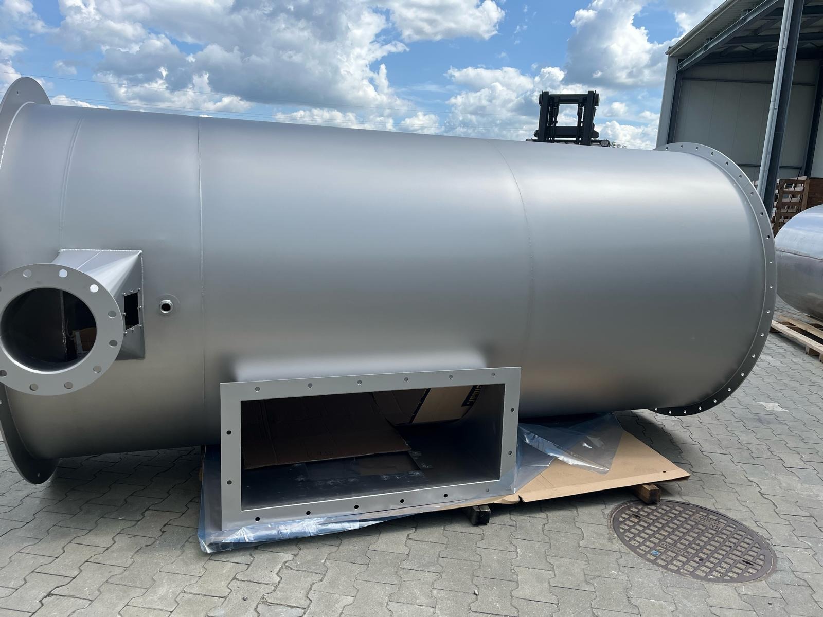 Szary, metalowy zbiornik przemysłowy z otworami i kołnierzami, leżący na zewnątrz hali. W tle widoczny wózek widłowy i fragment nieba.