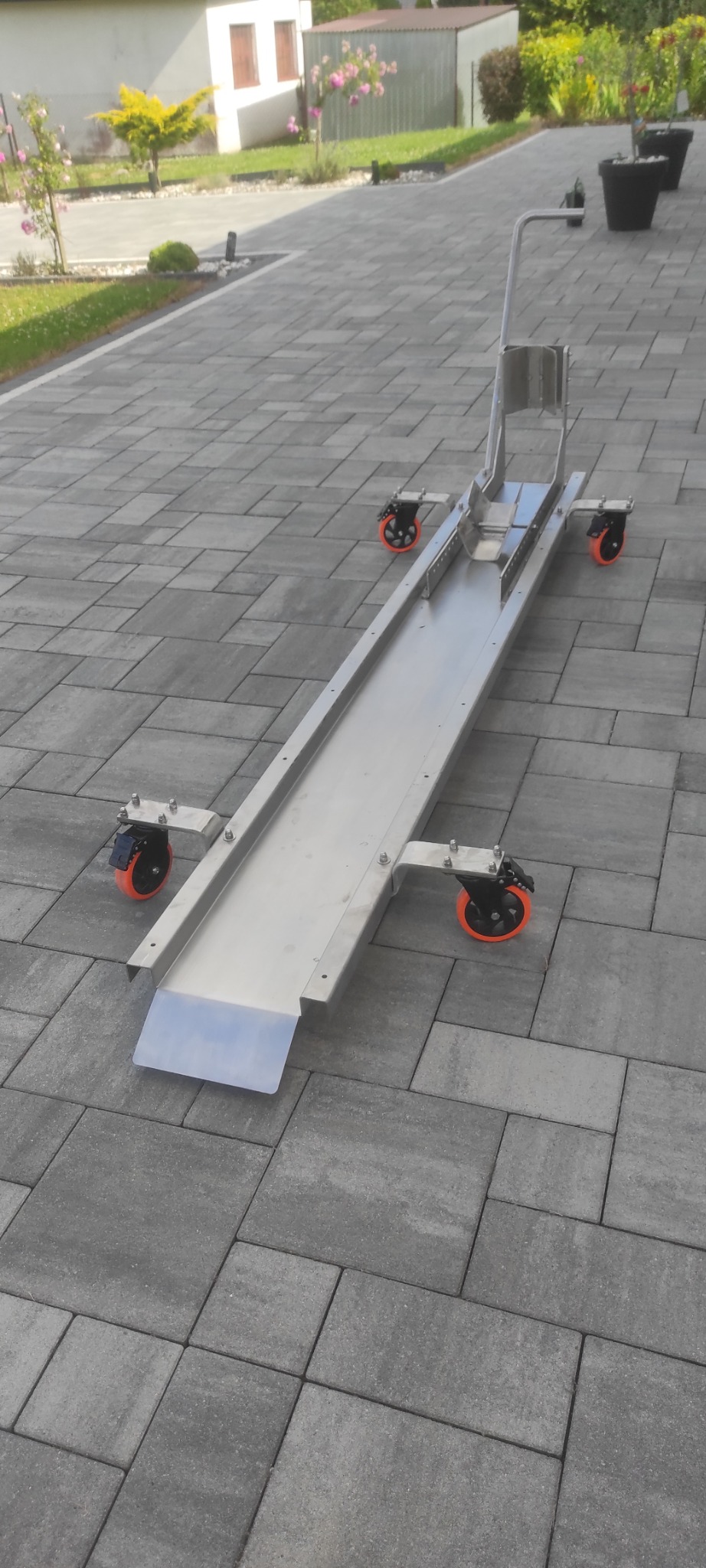 Wózek transportowy ze stali nierdzewnej na kółkach z pomarańczowymi felgami, ustawiony na szarej kostce brukowej. Widoczne elementy mocujące i uchwyt.