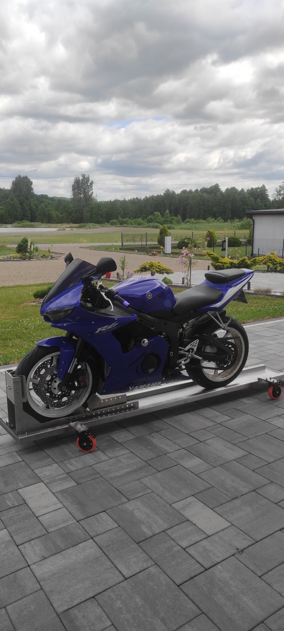 Niebieski motocykl Yamaha R6 na metalowej platformie z kółkami, na tle kostki brukowej i zieleni ogrodu. Widok z boku, pochmurne niebo.