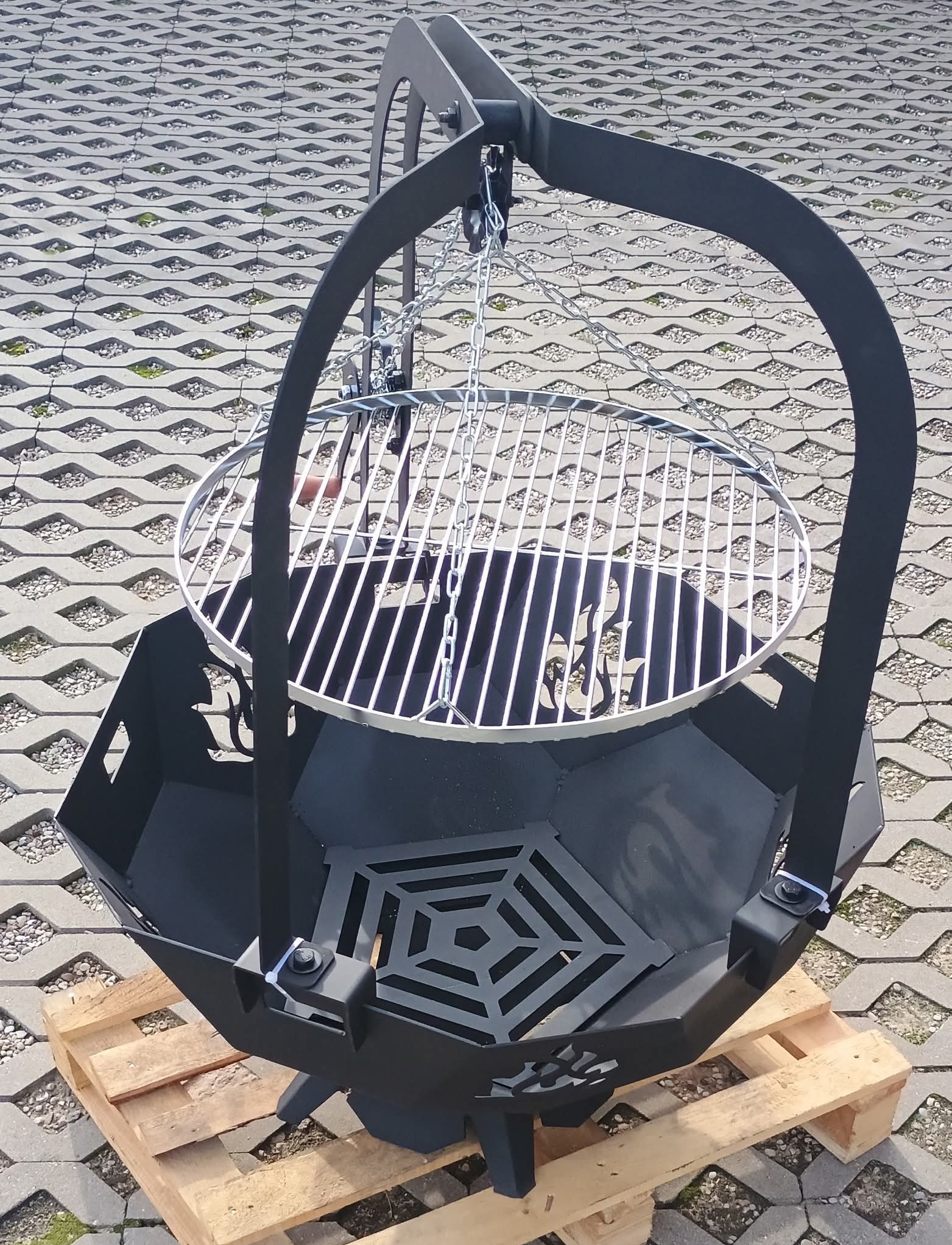 Metalowa konstrukcja paleniska z rusztem na łańcuchach, ustawiona na drewnianej palecie. Palenisko ma wycięte wzory i jest pomalowane na czarno.