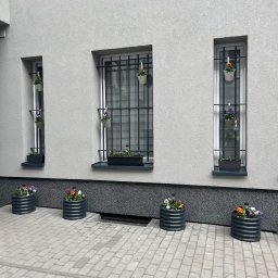 Dominik Hadryś - Szara fasada budynku z trzema oknami z kratami, ozdobionymi wiszącymi doniczkami. Na dole, wzdłuż ściany, ustawione są okrągłe, metalowe donice z kwiatami. Brukowana nawierzchnia.