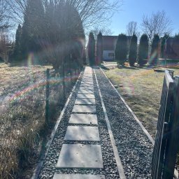 Dominik Hadryś - Ścieżka z płyt betonowych i grysu prowadząca przez ogród w słoneczny dzień. Widoczne drzewa i krzewy w tle. Elementy ogrodzenia po prawej stronie kadru.