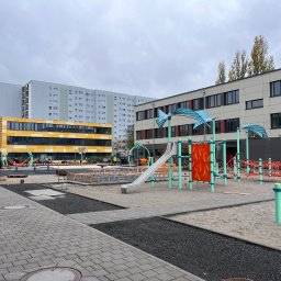 Dominik Hadryś - Plac zabaw w trakcie budowy: widoczne elementy konstrukcyjne, piasek, kostka brukowa i budynki w tle. Etap prac ziemnych i przygotowania podłoża.