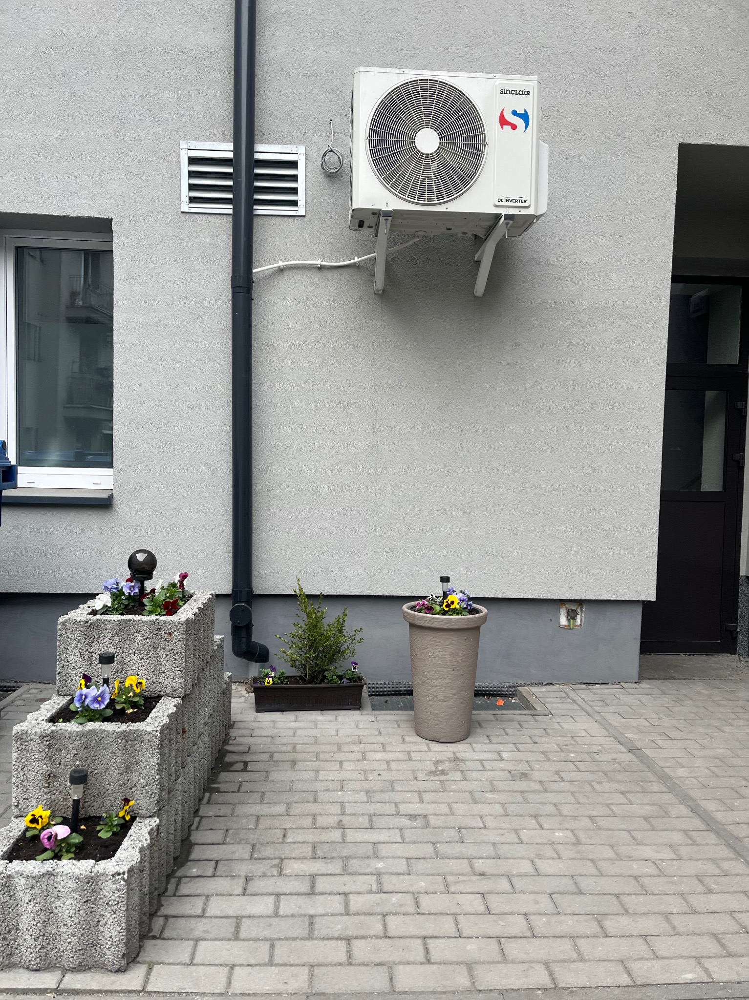 Szara elewacja budynku z klimatyzatorem Sinclair, oknem i donicami z kwiatami. Kostka brukowa na chodniku. Ujęcie z dołu, podkreślające detale.