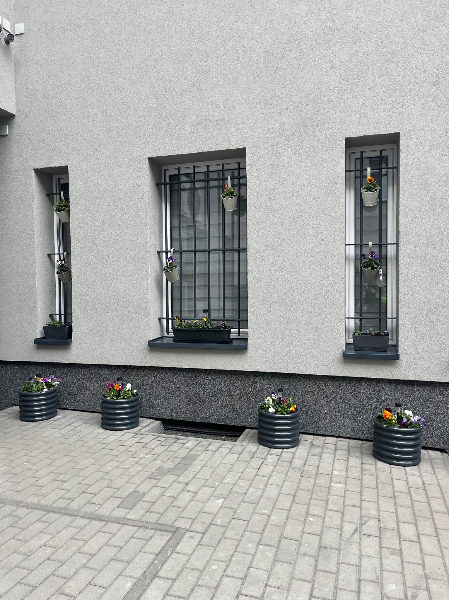 Szara fasada budynku z trzema oknami z kratami, ozdobionymi wiszącymi doniczkami. Na dole, wzdłuż ściany, ustawione są okrągłe, metalowe donice z kwiatami. Brukowana nawierzchnia.