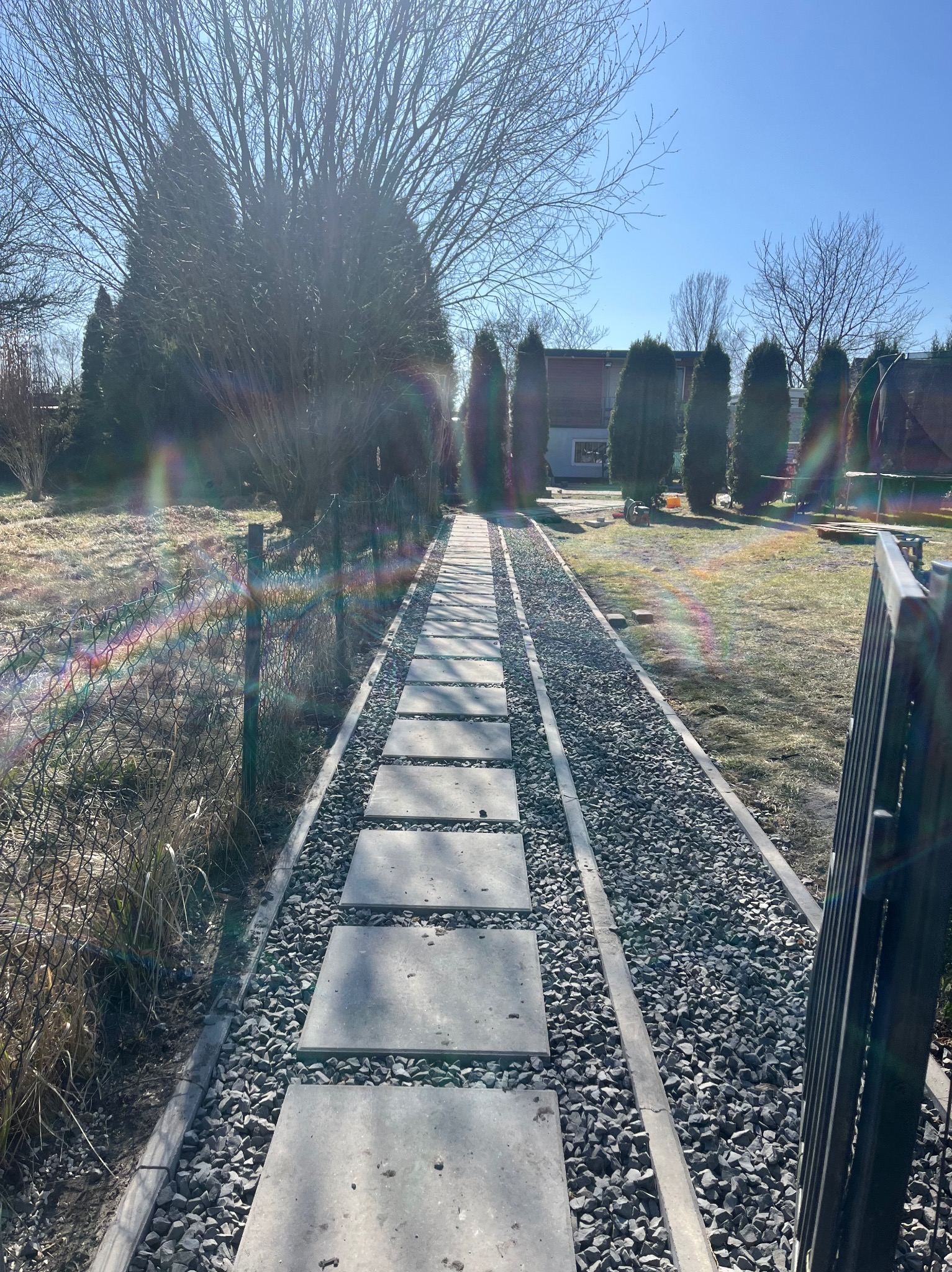 Ścieżka z płyt betonowych i grysu prowadząca przez ogród w słoneczny dzień. Widoczne drzewa i krzewy w tle. Elementy ogrodzenia po prawej stronie kadru.