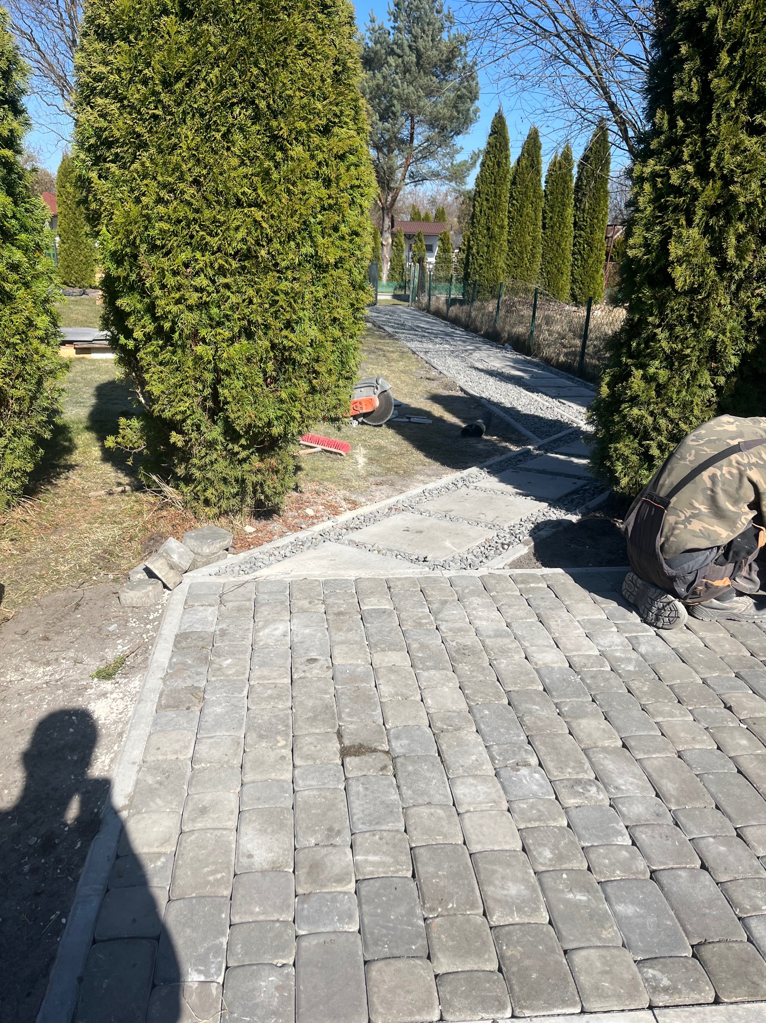 Układanie szarej kostki brukowej na ścieżce w ogrodzie w Katowicach. Widoczne narzędzia i fragment pracującego brukarza. Tuje w tle.