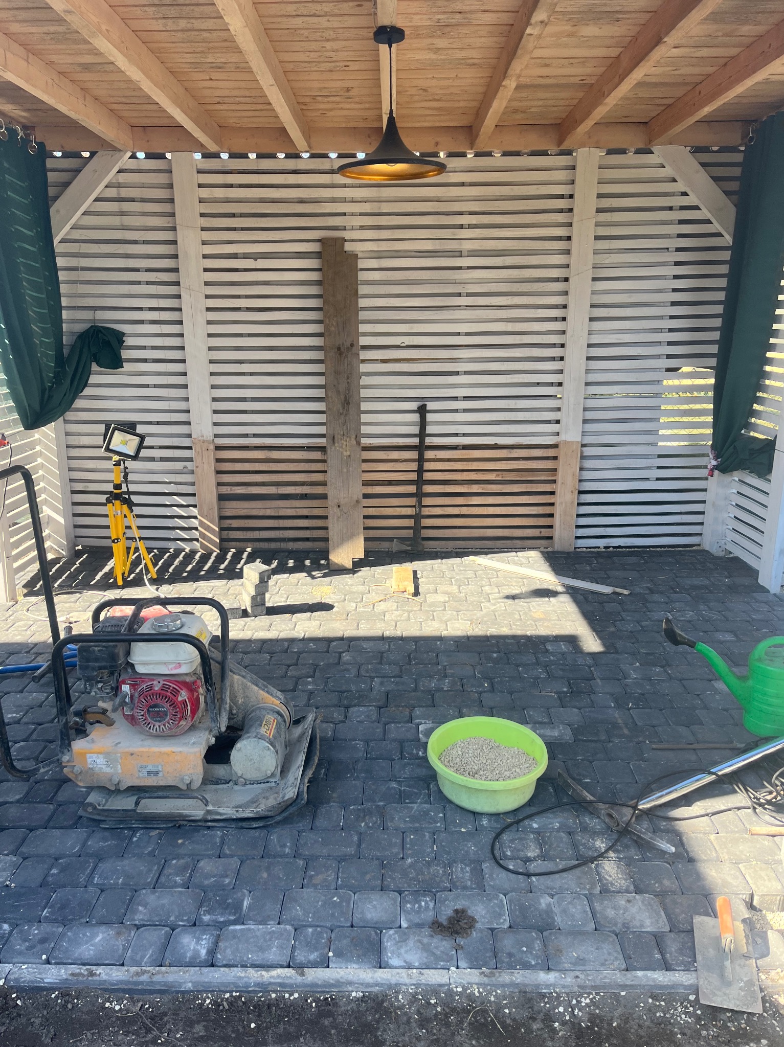 Częściowo ułożona, szara kostka brukowa pod zadaszeniem. Widoczna ubijarka, poziomica, wiadro z piaskiem i lampa na statywie. Prace brukarskie w toku.