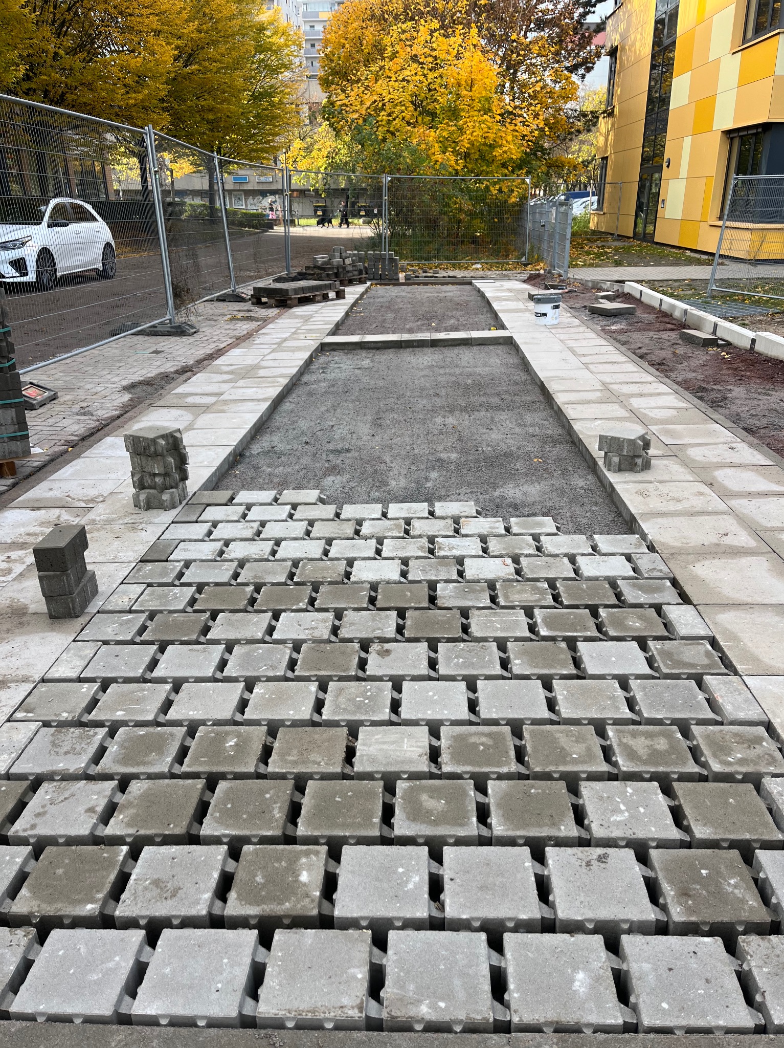 Fragment układanej ścieżki z betonowej kostki brukowej na przygotowanym podłożu. Widoczne stosy kostki, ogrodzenie i budynek w tle.