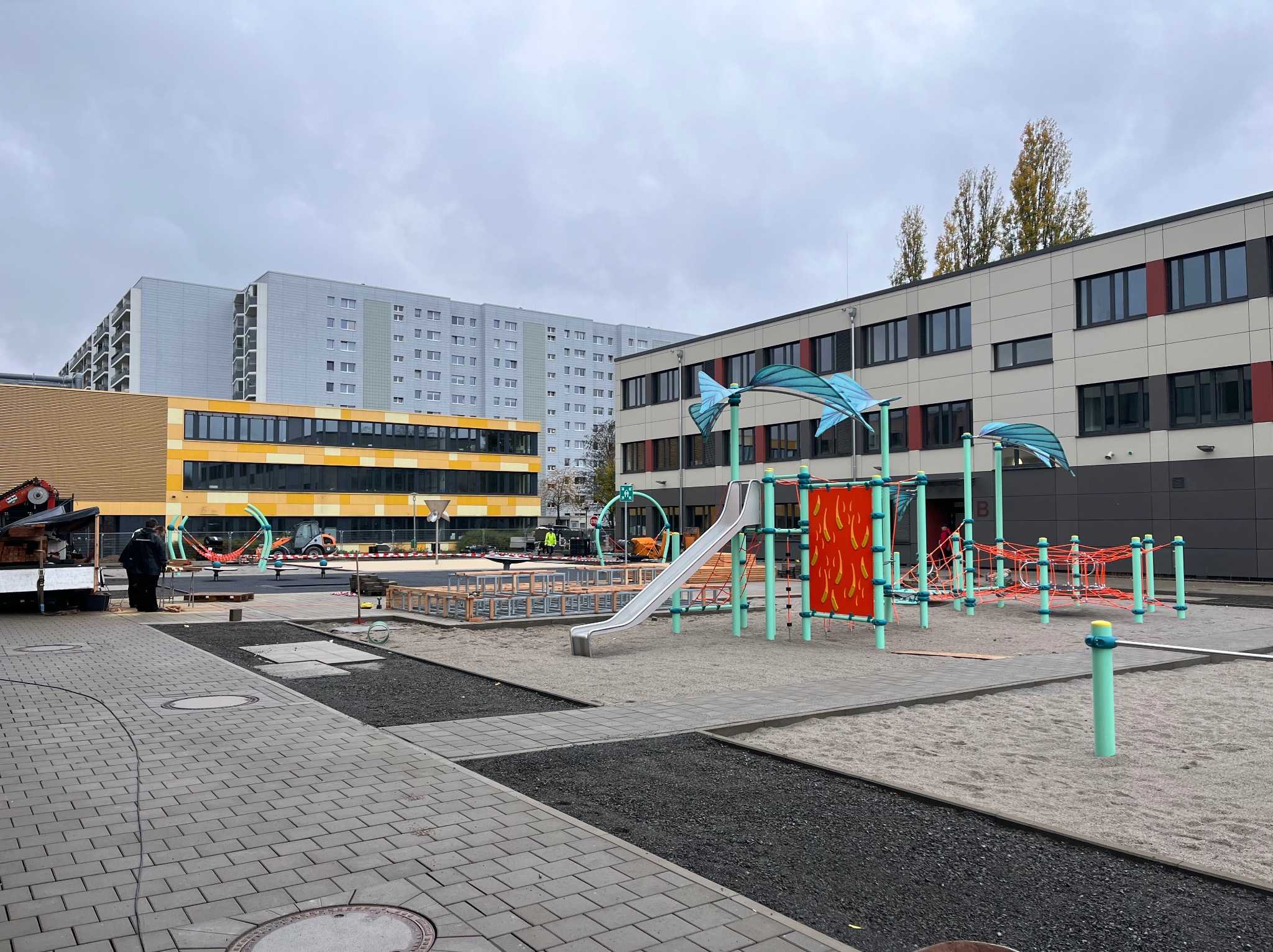 Plac zabaw w trakcie budowy: widoczne elementy konstrukcyjne, piasek, kostka brukowa i budynki w tle. Etap prac ziemnych i przygotowania podłoża.