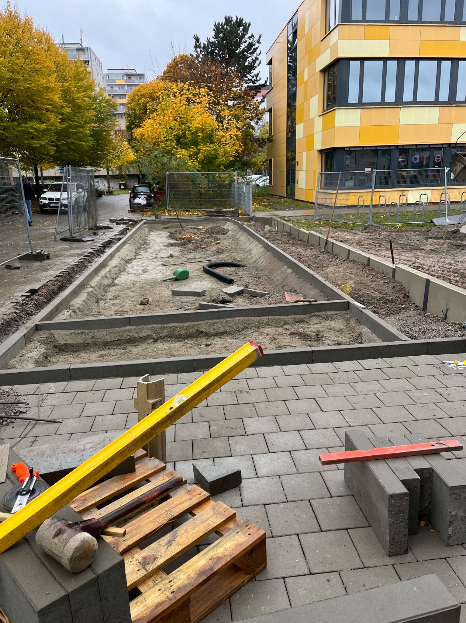 Etap przygotowania podłoża pod kostkę brukową z widocznymi narzędziami: poziomica, paleta, młotek. W tle budynek i drzewa w jesiennej scenerii.