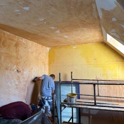 Wykończenia Premium WallPerfect - Dwóch mężczyzn szpachluje ścianę w pokoju na poddaszu. Widoczne rusztowanie, wiadro z farbą i narzędzia. Ściany w odcieniach żółci i brązu, częściowo pokryte szpachlą.