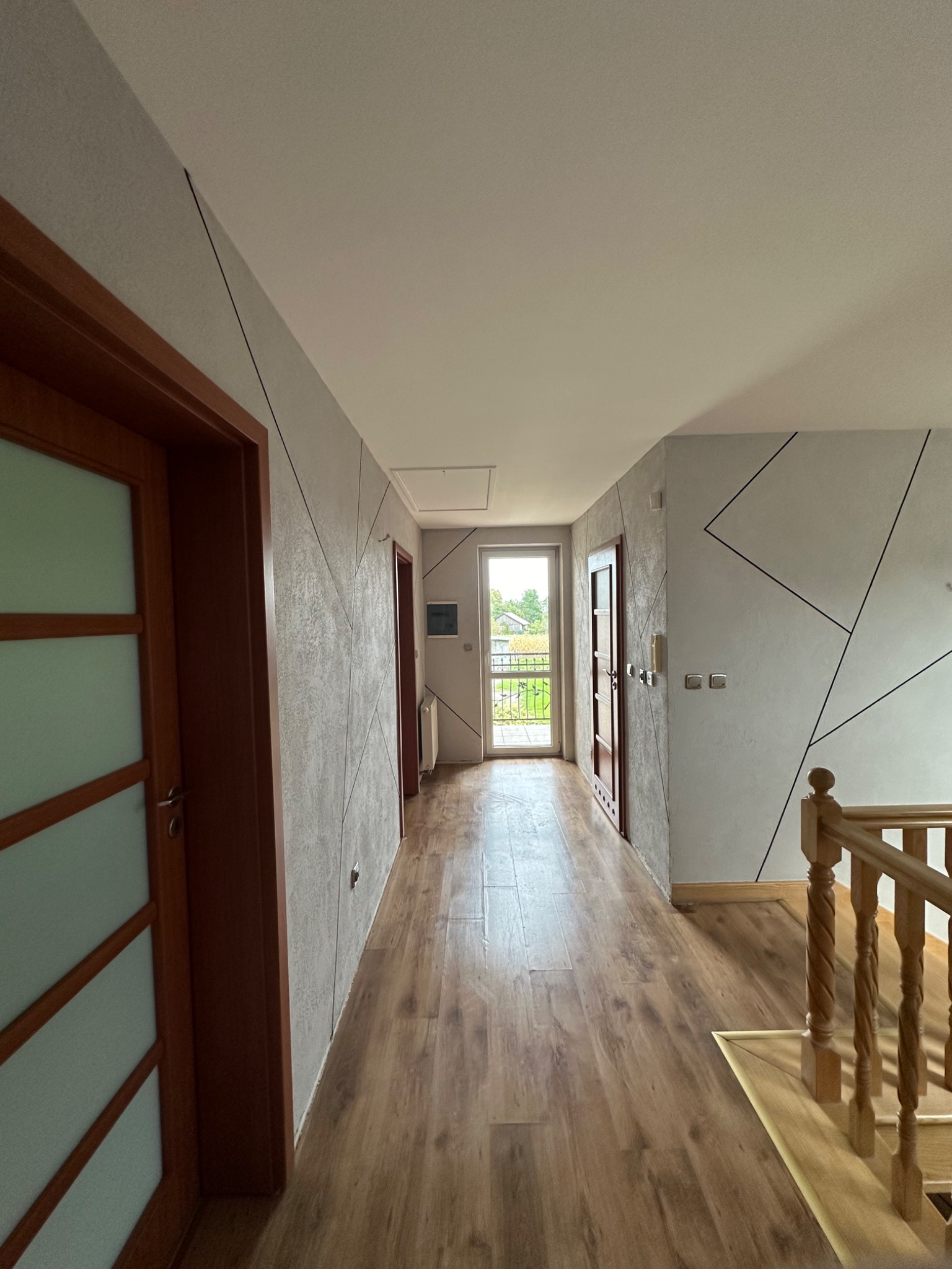 Korytarz po remoncie z drewnianą podłogą, szarymi ścianami z geometrycznym wzorem i drewnianymi drzwiami. Widok na balkon z zielenią w tle.
