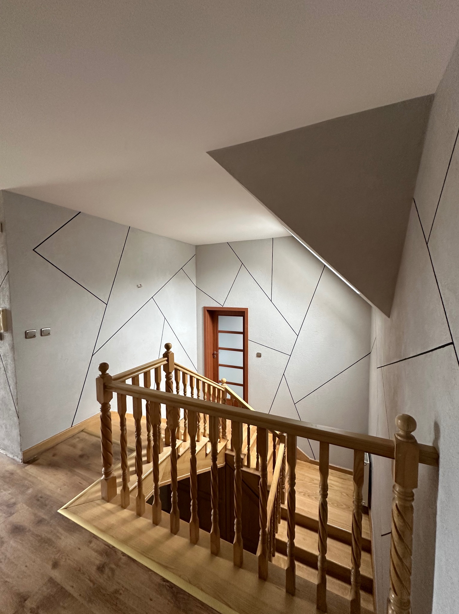 Klatka schodowa z drewnianymi schodami i balustradą. Ściany w jasnym odcieniu z geometrycznymi, czarnymi liniami. Widoczne drzwi i fragment sufitu.