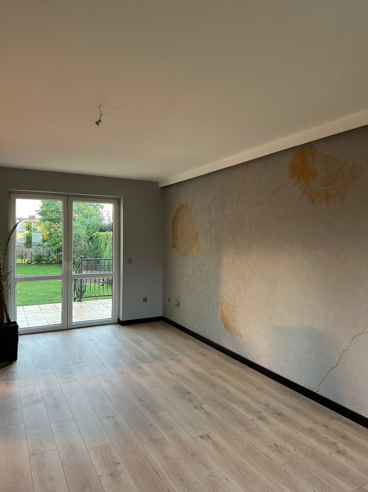 Jasne panele podłogowe i ściana z dekoracyjnym, złotym wzorem w przestronnym pokoju z wyjściem na balkon i ogród. Widok na zieleń za oknem.