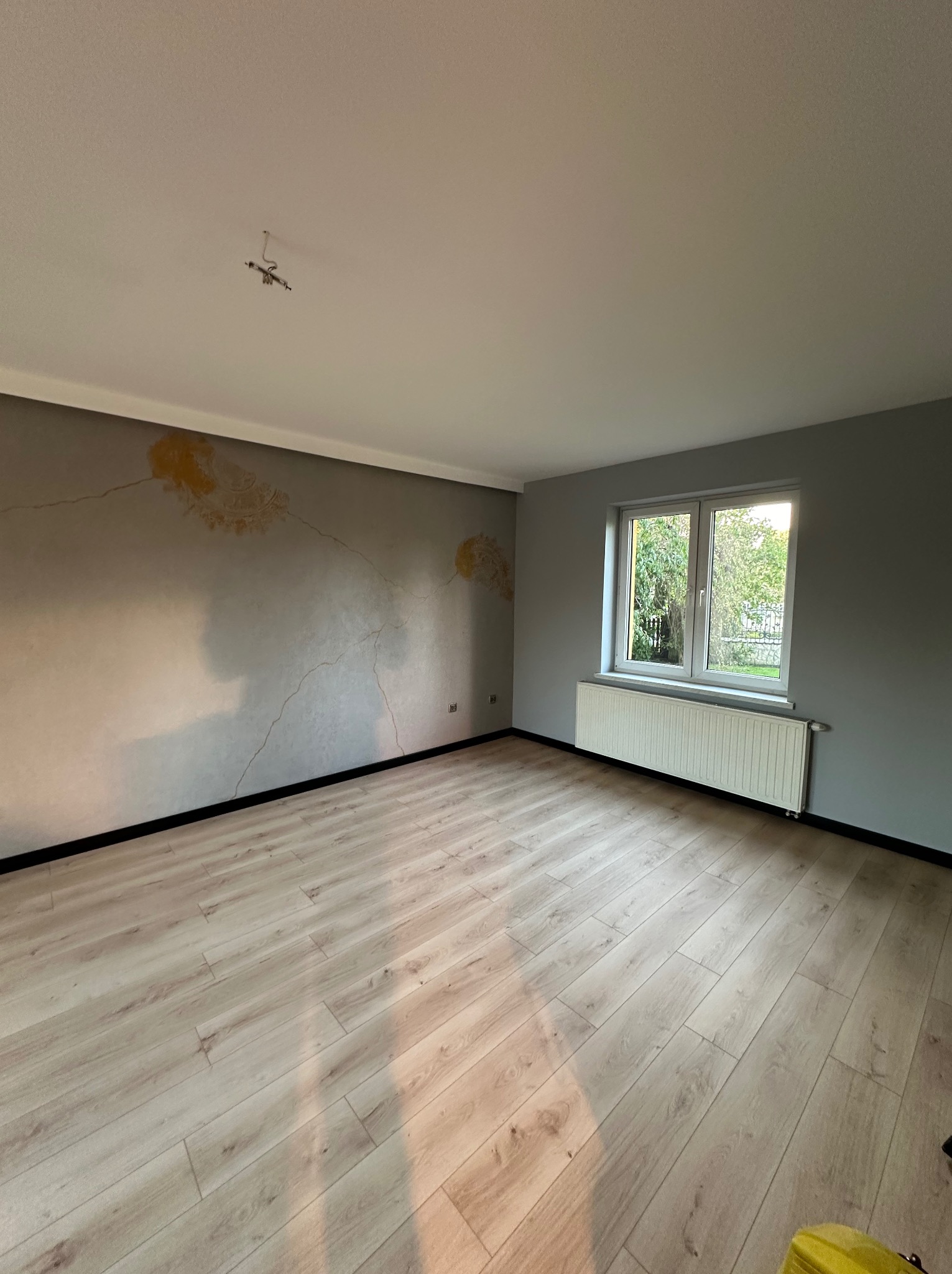 Jasne panele podłogowe w pustym pokoju po malowaniu ścian na szaro z dekoracyjnym wzorem. Okno z białym grzejnikiem. Widoczne niedokończone podłączenie lampy.