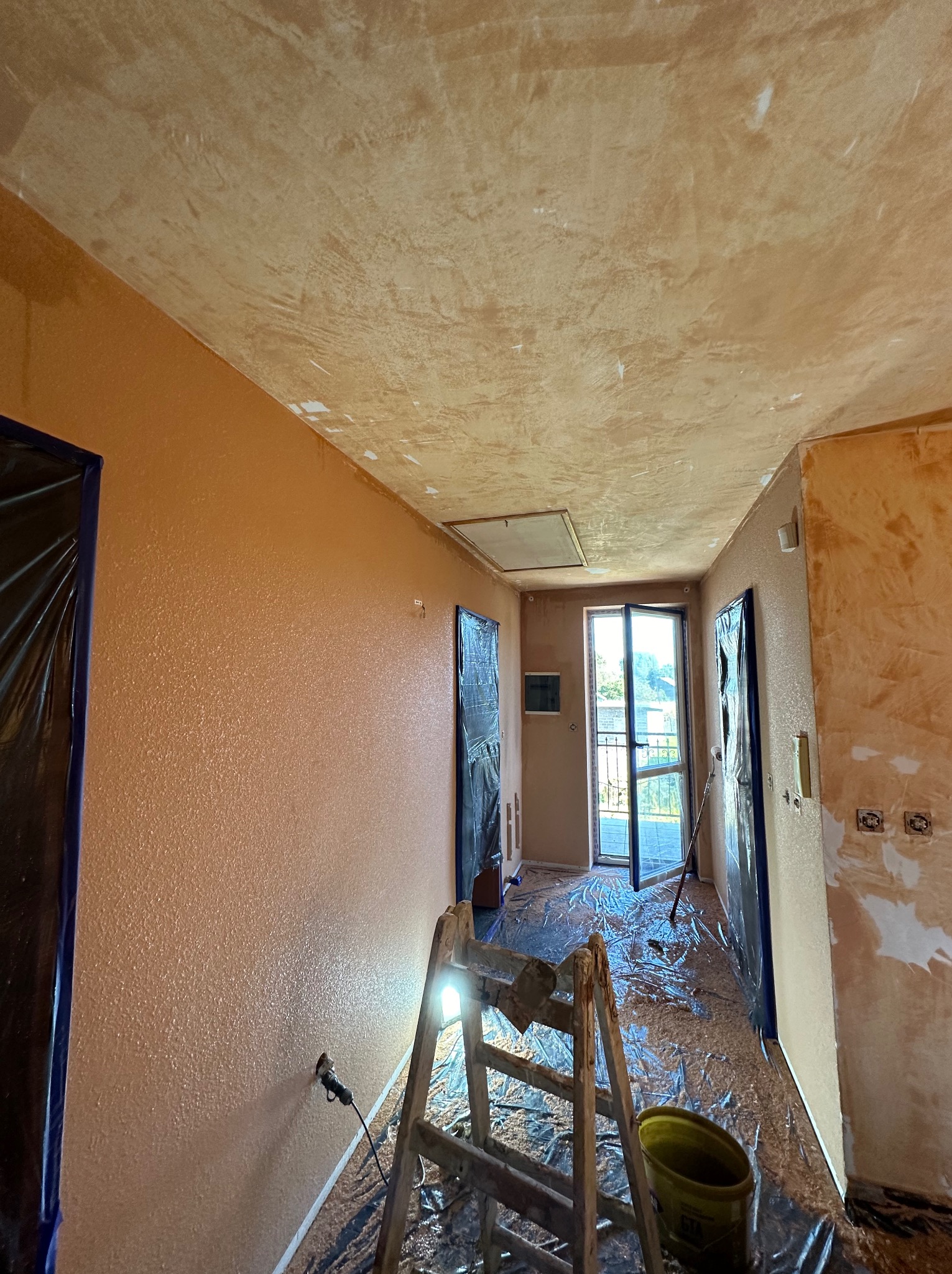 Pokój po malowaniu w Skomielnej Czarnej z zabezpieczonymi drzwiami folią. Widoczna drabina i wiadro. Ściany w odcieniach beżu i brązu, sufit z efektem dekoracyjnym.