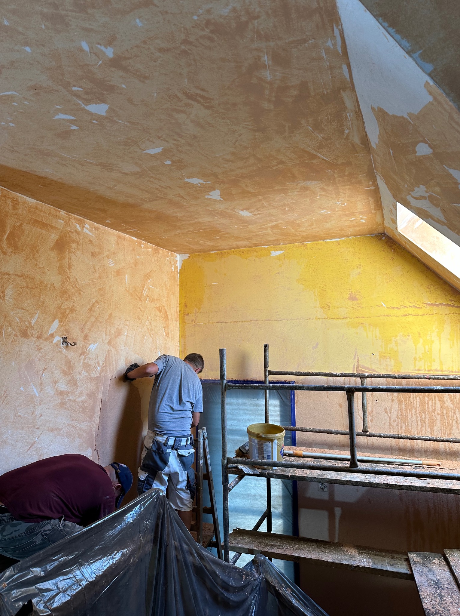 Dwóch mężczyzn szpachluje ścianę w pokoju na poddaszu. Widoczne rusztowanie, wiadro z farbą i narzędzia. Ściany w odcieniach żółci i brązu, częściowo pokryte szpachlą.