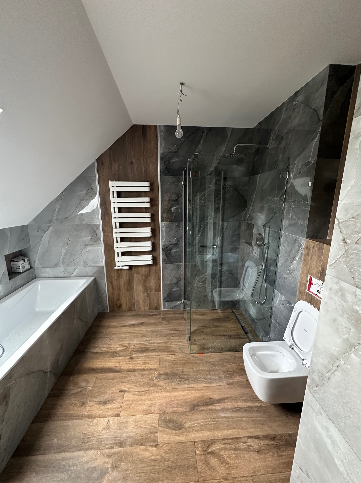 Nowoczesna łazienka na poddaszu z kabiną prysznicową, wiszącą toaletą, grzejnikiem drabinkowym i wanną. Płytki imitujące marmur i drewno. Ukośny sufit.