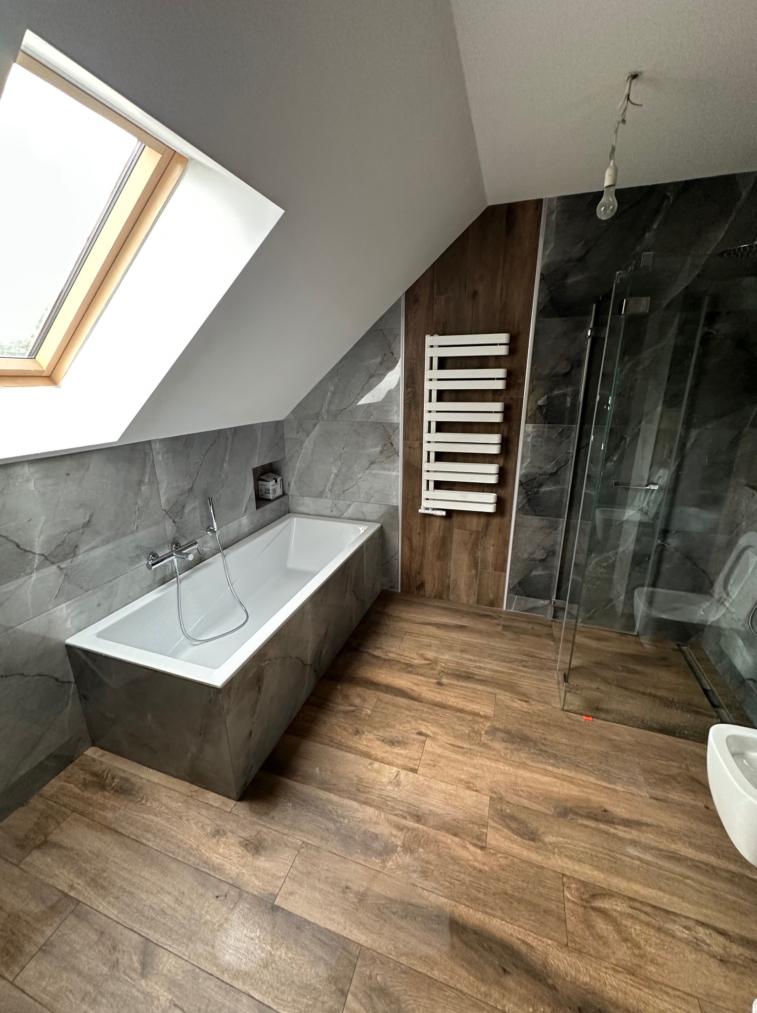 Nowoczesna łazienka na poddaszu z oknem dachowym, wykończona płytkami imitującymi marmur i drewno. Kabina prysznicowa ze szklaną ścianą, grzejnik drabinkowy.