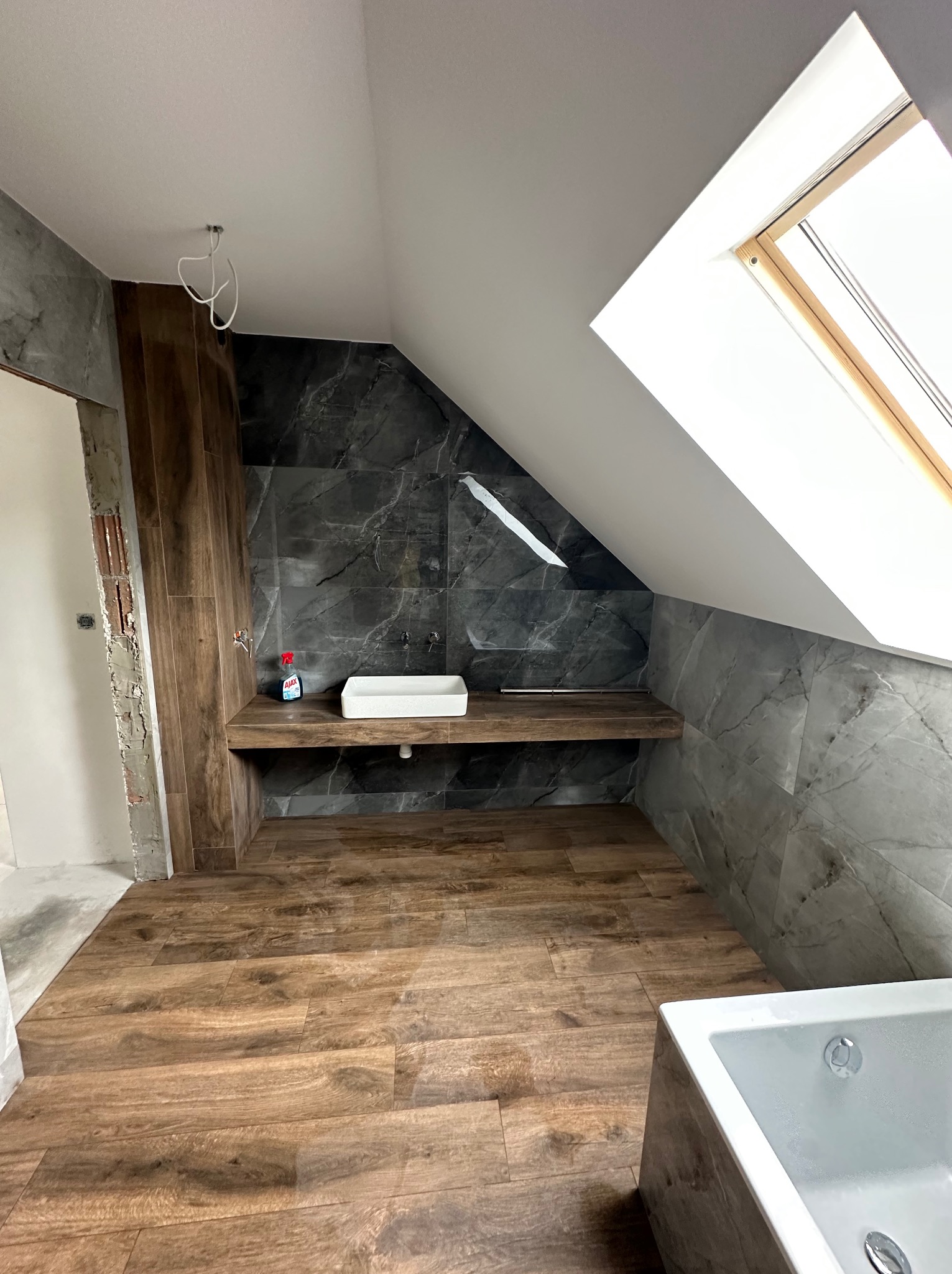 Łazienka na poddaszu z drewnopodobną podłogą i szarą marmurową ścianą. Umywalka na drewnianym blacie, okno dachowe. Częściowo wykończone ściany.