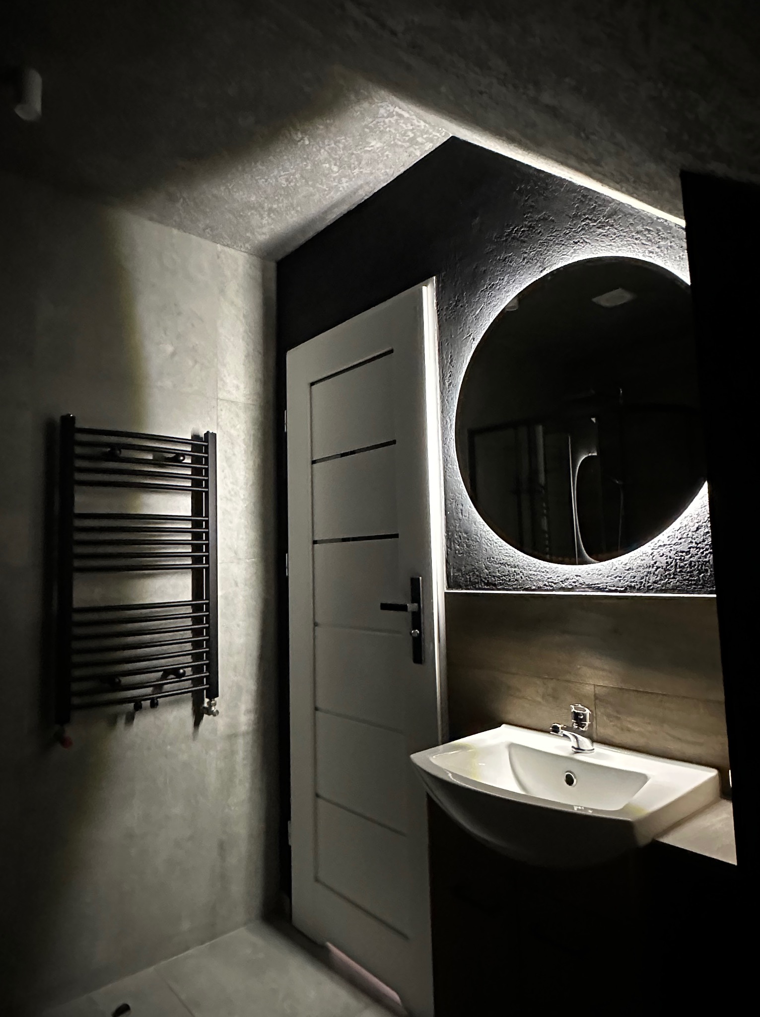 Nowoczesna łazienka z okrągłym lustrem z podświetleniem LED, umywalką, grzejnikiem drabinkowym i białą, minimalistyczną stolarką drzwiową. Ciemna tonacja wnętrza.