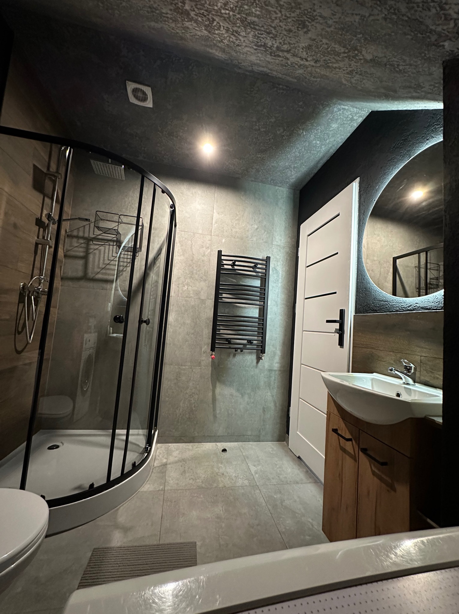 Nowoczesna łazienka z kabiną prysznicową typu walk-in, umywalką na drewnianej szafce i okrągłym lustrem z podświetleniem. Ciemna kolorystyka ścian i sufitu.