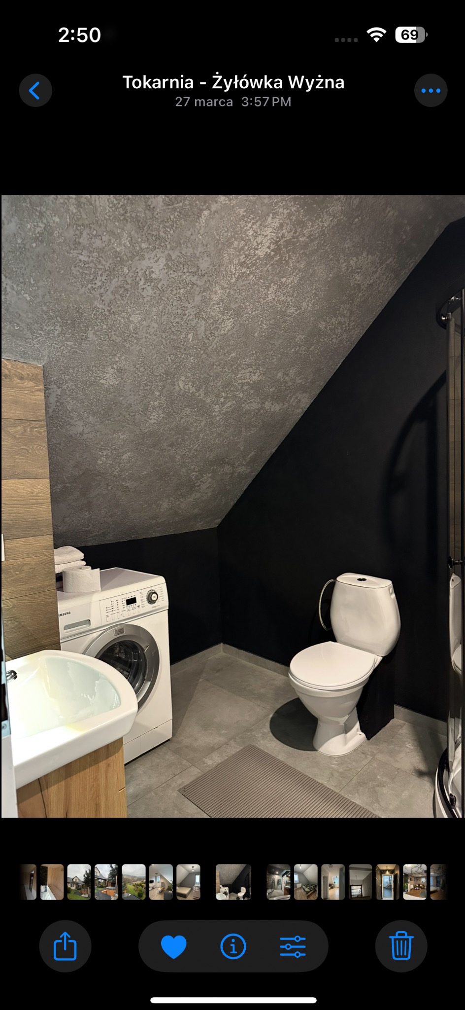 Nowoczesna łazienka na poddaszu z pralką, umywalką i toaletą. Ściany pomalowane na czarno, sufit w odcieniach szarości. Minimalistyczny design, szare płytki podłogowe.