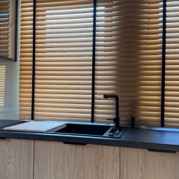 KAPRYS - Kuchnia z drewnianymi żaluzjami poziomymi w ciepłym odcieniu, czarne akcenty ramy okiennej i armatury, nowoczesny design, naturalne światło.