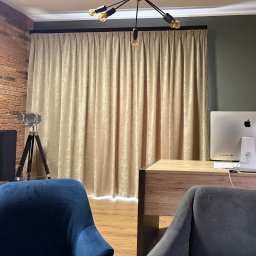 KAPRYS - Beżowe, gładkie zasłony w salonie z ceglaną ścianą i nowoczesną lampą sufitową. Widoczny fragment biurka z komputerem i krzesła.