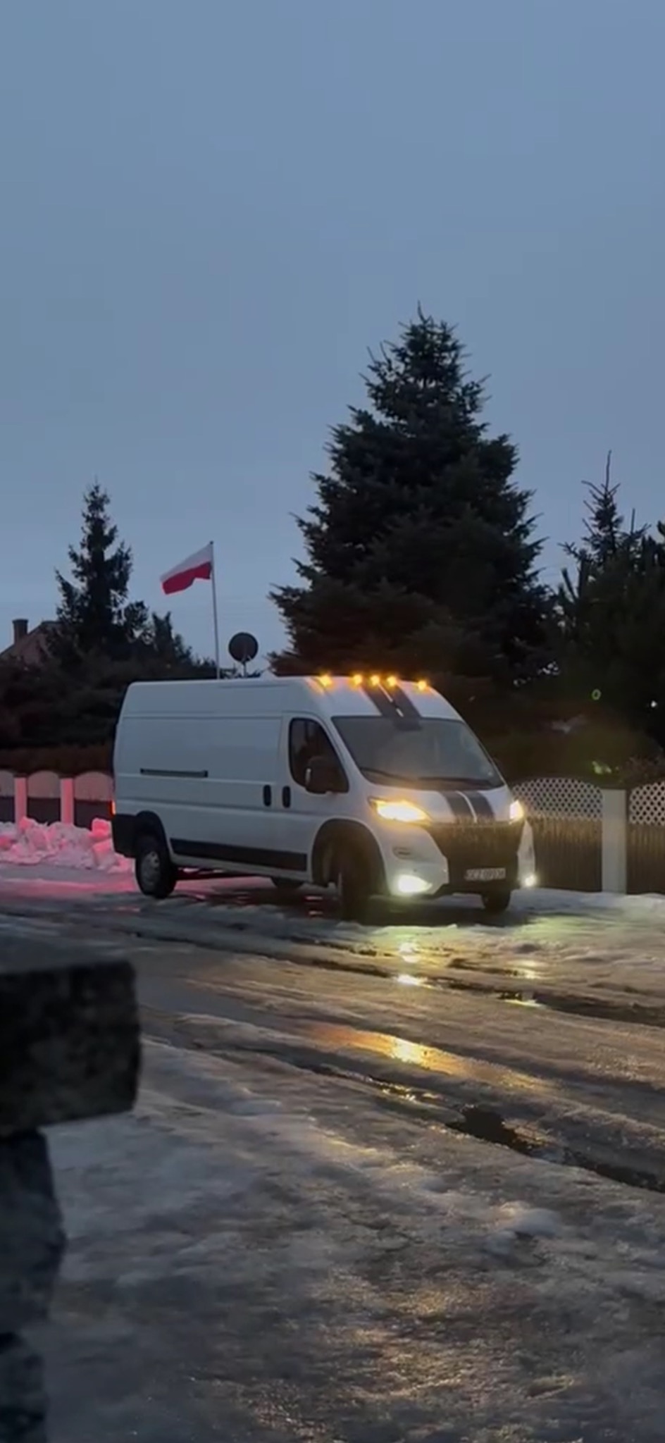 Biały bus z czarnymi pasami na dachu i polską flagą, zaparkowany na ośnieżonej drodze w zimowy wieczór. Włączone światła oświetlają drogę.