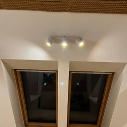 Józef Bujakowski - Biała lampa sufitowa z trzema reflektorami oświetlająca dwa okna dachowe w skosie, z dekoracyjnym sznurem na suficie. Ciepłe światło.