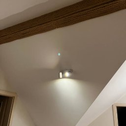 Józef Bujakowski - Zamontowana, nowoczesna lampa sufitowa z dwoma reflektorami, oświetlająca białe wnętrze z dekoracyjnym elementem z liny na suficie. Minimalistyczny design.