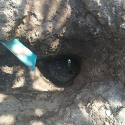 Józef Bujakowski - Wykop w ziemi z wystającą rurką instalacyjną i fragmentem niebieskiej taśmy ostrzegawczej. Widok z góry na przygotowany otwór pod instalację.