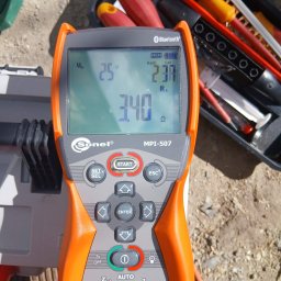 Józef Bujakowski - Miernik Sonel MPI-507 z wyświetlonymi wartościami pomiarów elektrycznych, obok skrzynka z narzędziami, na tle ziemi. Pomiar napięcia i rezystancji.