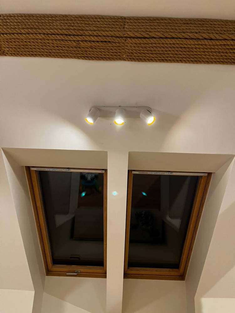 Biała lampa sufitowa z trzema reflektorami oświetlająca dwa okna dachowe w skosie, z dekoracyjnym sznurem na suficie. Ciepłe światło.