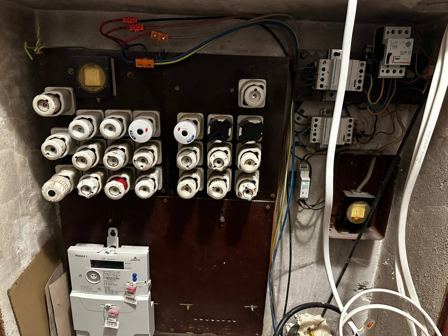 Stara tablica rozdzielcza z bezpiecznikami topikowymi, licznikiem energii i przewodami. Widoczne elementy instalacji elektrycznej wewnątrz budynku.